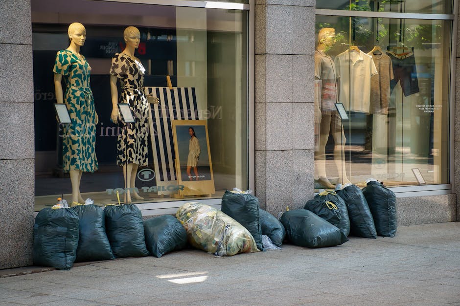 A row of dark green rubbish bags, some tied tightly and others partially torn open, are lined up along the exterior of a clothing store window on a city sidewalk. The bags appear filled with discarded