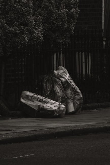 Two large black plastic garbage bags, filled and tied at the top, are leaning against a low wooden fence on a paved sidewalk. The bags appear to contain household waste or debris and are situated near