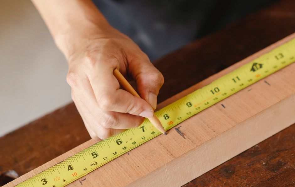 A close-up of a person's hand measuring a piece of light-colored wood with a yellow measuring tape that displays both imperial and metric units, with the hand holding a pencil at the 8-inch mark. The
